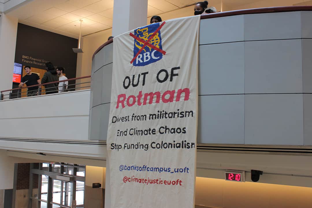 Banner hanging off a ledge in the Rotman School of Management building, showing a red cross over the RBC logo with the main text stating "Out of Rotman".